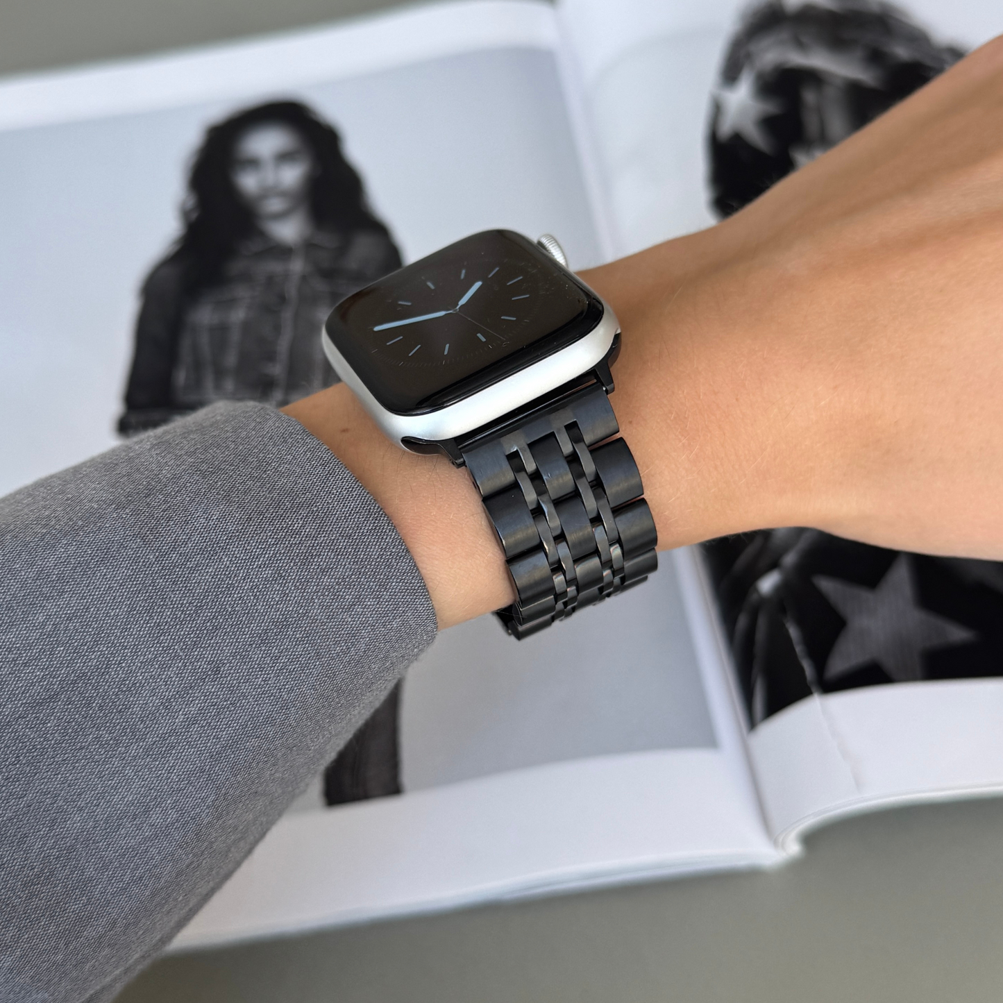 Black link Apple Watch band worn with a grey sleeve over a magazine spread. Waterproof stainless steel iWatch bracelet, compatible with Apple Watch 38mm to 49mm, Series 1-11, SE and Ultra