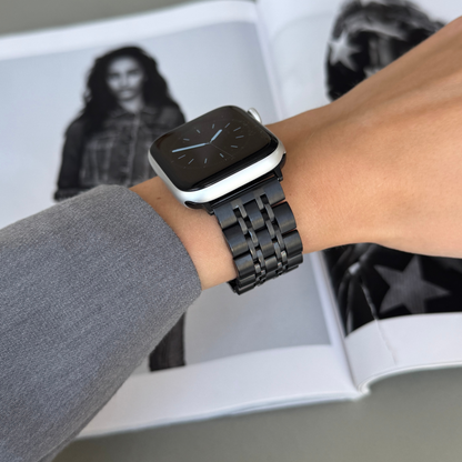 Black link Apple Watch band worn with a grey sleeve over a magazine spread. Waterproof stainless steel iWatch bracelet, compatible with Apple Watch 38mm to 49mm, Series 1-11, SE and Ultra