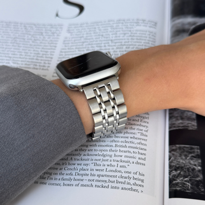 Silver link Apple Watch band worn on a wrist above an open magazine. Minimalist stainless steel iWatch strap, compatible with Apple Watch 38mm to 49mm, Series 1-11, SE and Ultra
