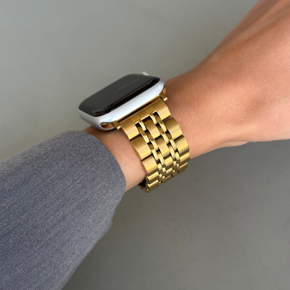 Gold link Apple Watch band worn on a wrist with a grey sleeve. Waterproof stainless steel metal armband, compatible with Apple Watch 38mm to 49mm, Series 1-11, SE and Ultra