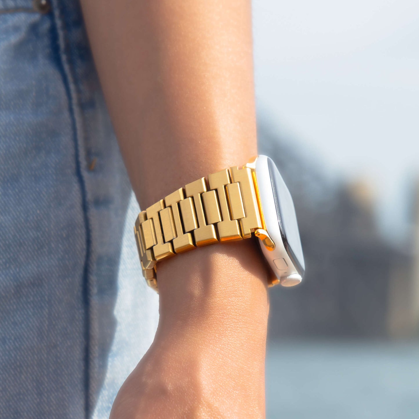Gold link Apple Watch band worn on a wrist over denim outdoors. Sleek unisex stainless steel iWatch bracelet with a brushed finish, compatible with Apple Watch 38mm, 40mm, 41mm, 42mm, 44mm, 45mm, 46mm and 49mm, Series 1-11, SE and Ultra