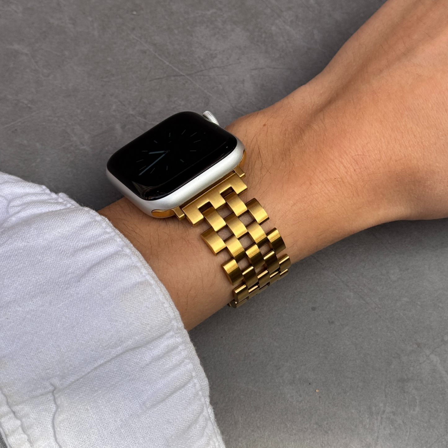 Gold multi link Apple Watch band worn on a wrist against a grey wall. Modern unisex stainless steel iWatch strap with clean geometry, compatible with Apple Watch 38mm to 49mm, Series 1-11, SE and Ultra