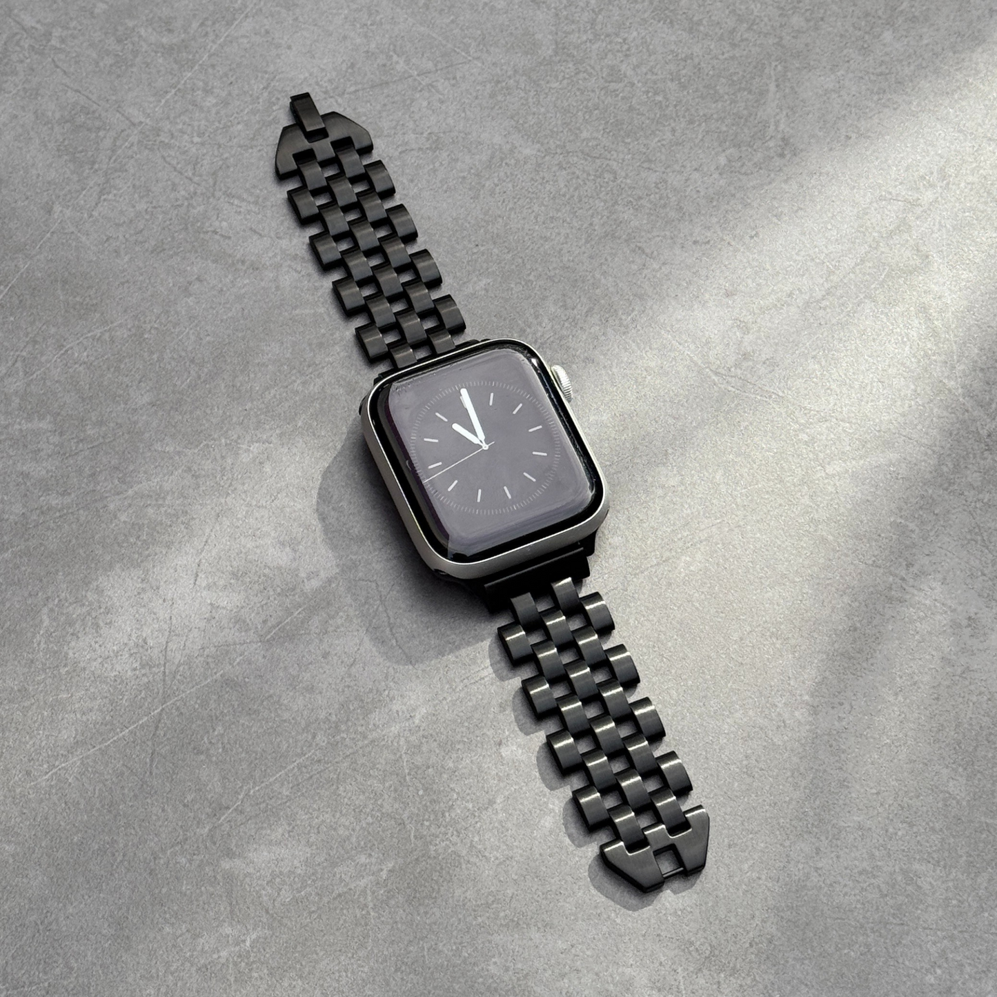 Black multi link Apple Watch band lying flat on grey concrete. Modern stainless steel iWatch strap with a seamless link profile, compatible with Apple Watch 38mm to 49mm, Series 1-11, SE and Ultra
