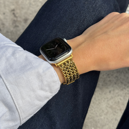 A close-up of a person's wrist wearing a gold tone mesh metal band on a Apple smartwatch, with dark blue jeans and a light-colored shirt visible in the background