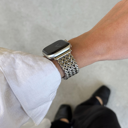 A close-up of a person's wrist wearing a silver metal mesh watch band with an Apple Watch. The person is wearing a white long-sleeve shirt and black pants, standing on a light gray concrete floor.