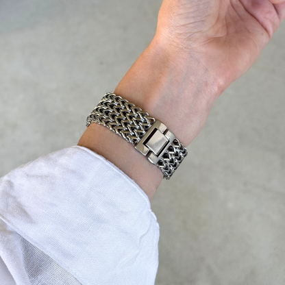 A close-up of a silver toned metal link bracelet with a wide, textured woven-style Apple Watch band and a rectangular clasp, worn on a person's wrist with a white sleeve visible.