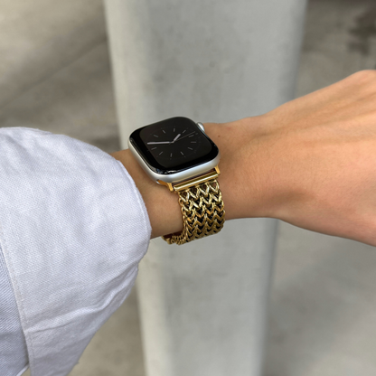 A close-up of a person's wrist wearing a gold tone metal mesh watch band on an Apple Watch, with a light blue shirt sleeve visible and a blurred gray concrete background.