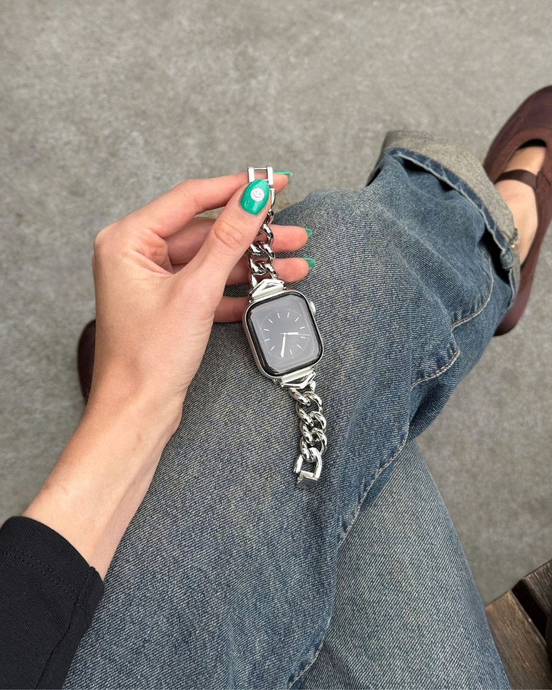 A silver metal chain Apple Watch band is held by a hand with green nail polish, resting on a person's denim-clad leg against a gray floor.