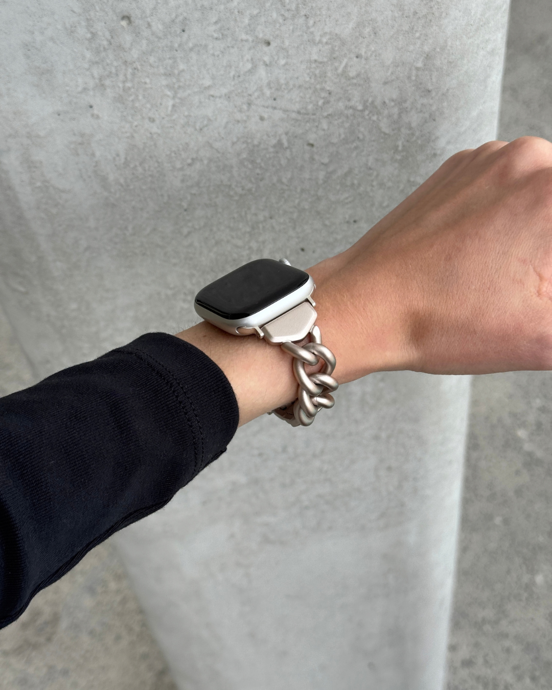 A close-up of a person's wrist wearing a chunky starlight chain link watch strap on an Apple Watch, set against a gray concrete pillar background.