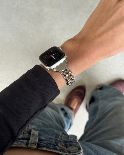 A close-up lifestyle shot of a person wearing a silver toned chunky chain link apple watch band. The person is also wearing a black long-sleeve top, blue jeans, and brown shoes, standing on a light gray floor.