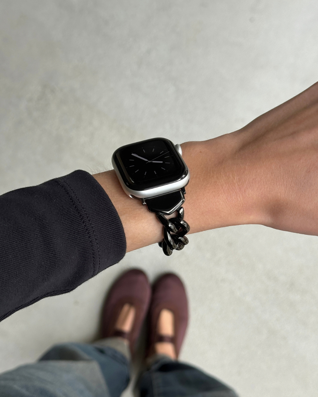 A black chain-link smartwatch band worn on a person's wrist, with an Apple Watch 11. The person is wearing a dark long-sleeve top, jeans, and burgundy Mary Jane flats, photographed from a top-down perspective against a light gray concrete floor.