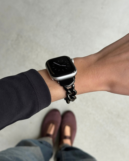 A black chain-link smartwatch band worn on a person's wrist, with an Apple Watch 11. The person is wearing a dark long-sleeve top, jeans, and burgundy Mary Jane flats, photographed from a top-down perspective against a light gray concrete floor.