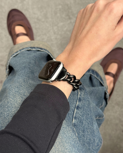 A close-up lifestyle shot of a person wearing a chunky black chain-link Apple Watch band paired with blue jeans, a black long-sleeve top, and brown Mary Jane shoes.