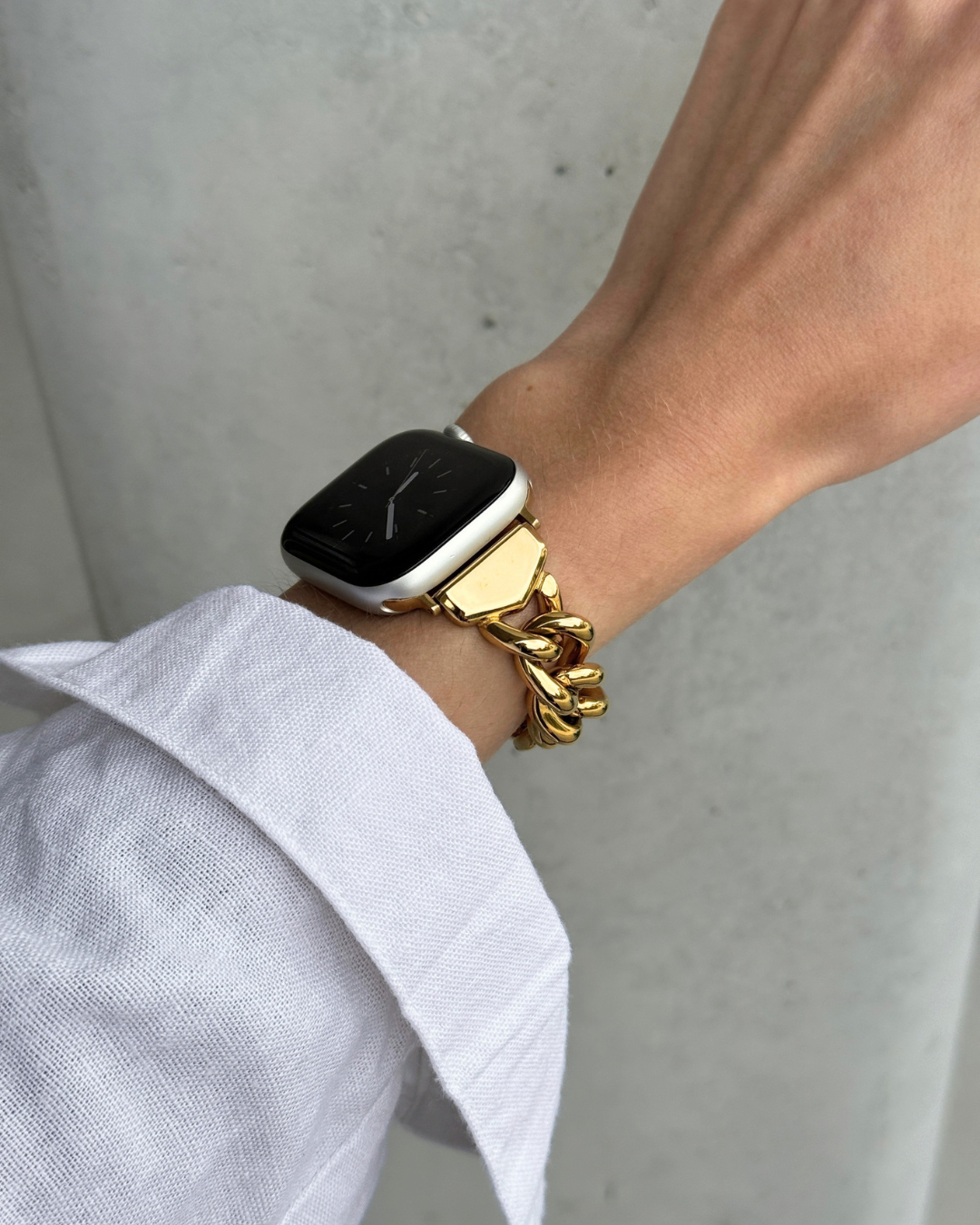 A close-up of a person's wrist wearing a chunky gold chain link bracelet with an Apple Watch, paired with a white collared shirt against a gray concrete wall.