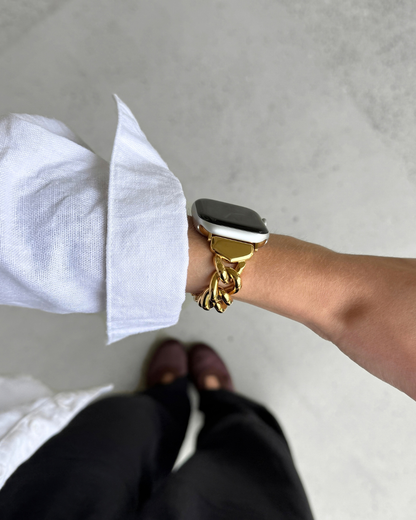 A close-up of a person's wrist wearing a chunky gold chain bracelet with an Apple watch, paired with a white shirt and dark pants, photographed from a top-down perspective.