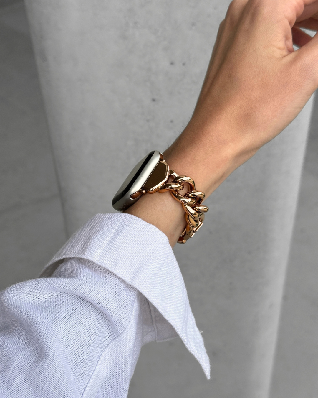 A close-up of a person's wrist wearing a chunky rose gold chain bracelet on an Apple Watch, with a white linen shirt cuff visible against a gray concrete background.