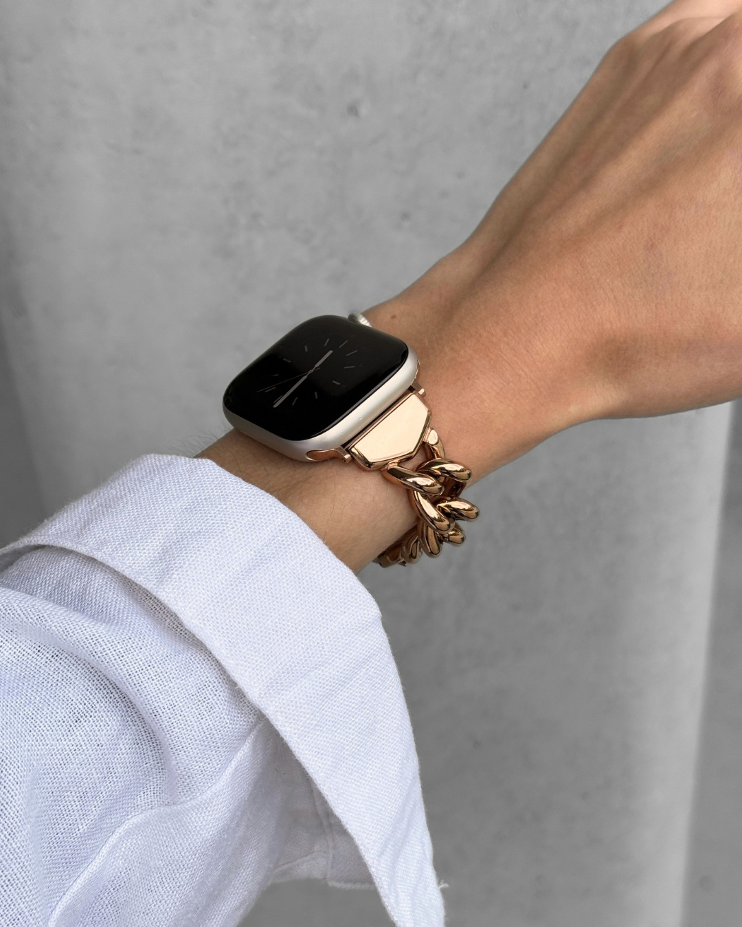 A close-up of a person's wrist wearing a rose gold toned chain Apple Watch Band, paired with a white shirt cuff against a gray concrete wall.