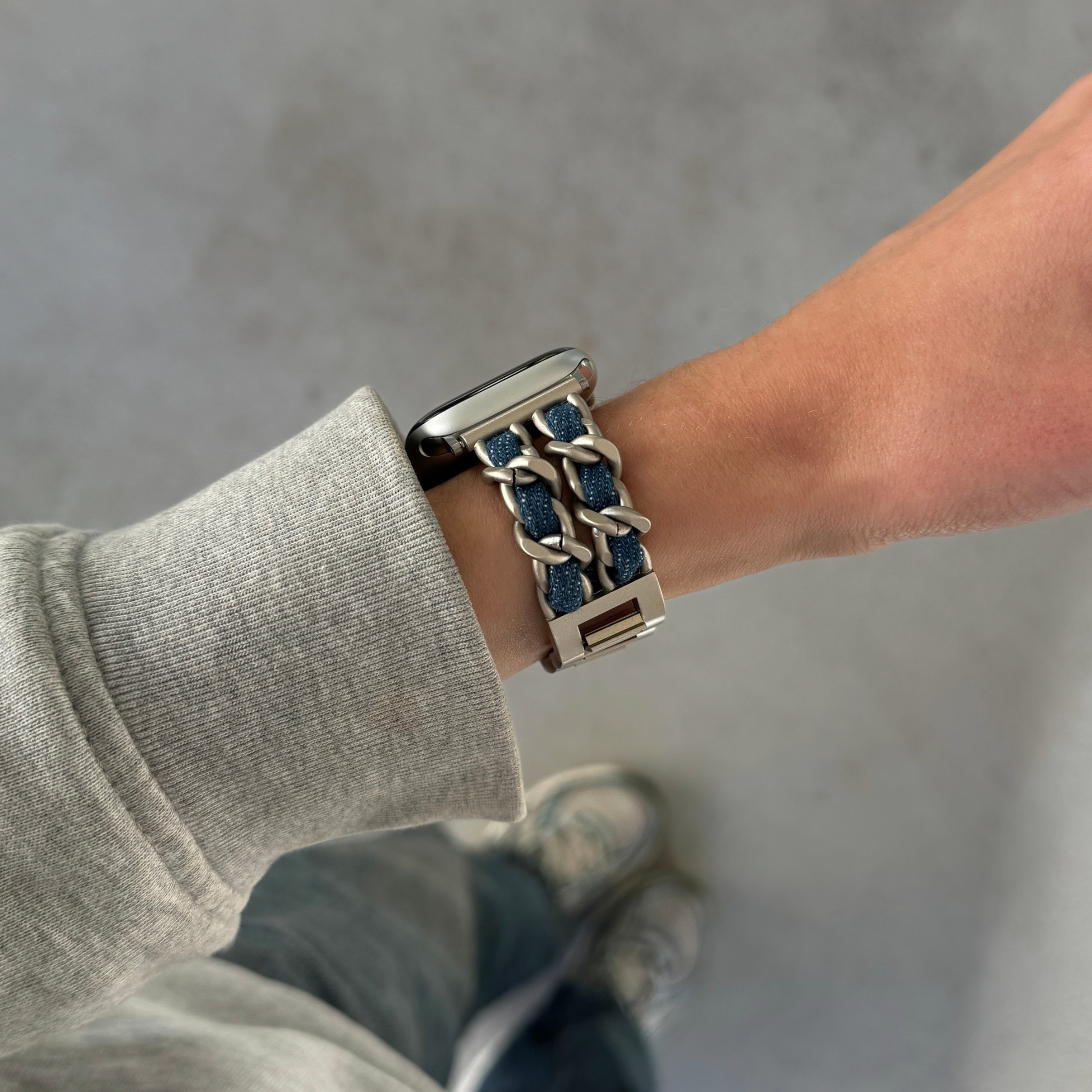A close-up of a person's wrist wearing a starlight toned chain-link Apple watch band with textured denim inlay, paired with a ribbed sweatshirt and jeans against a concrete floor.